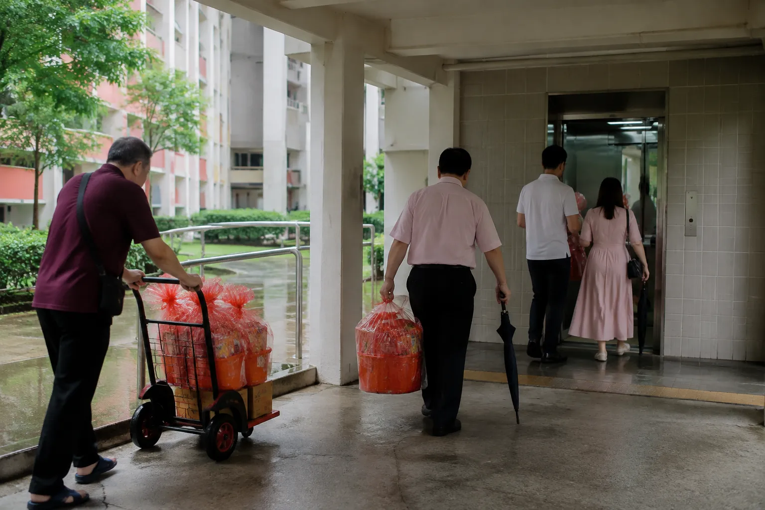 Labelled wedding gift packages being carried through sheltered access toward an HDB lift